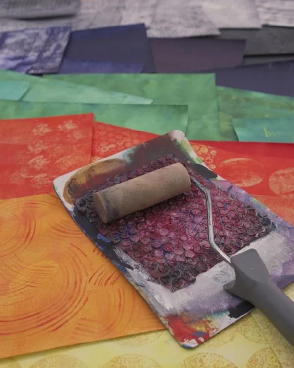 Nahaufnahme eines Farbrollers, der mit roter Farbe bedeckt ist und auf einer flachen, rechteckigen Palette mit rosa und violetter Farbe liegt. Die Palette liegt auf einer Reihe von quadratischen, farbigen Papierstücken, die in einem Regenbogenmuster angeordnet sind, mit roten, orangefarbenen, gelben, grünen und blauen Farbtönen.