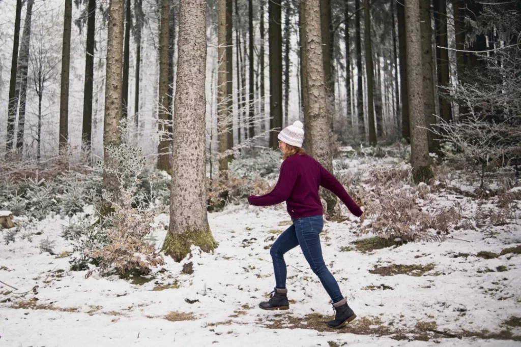 Eine Frau mit einer rosa Mütze und einem weinroten Pullover geht in einem verschneiten Wald spazieren. Sie trägt blaue Jeans und schwarze Stiefel. Die Bäume im Wald sind hoch und schlank, und der Boden ist mit Schnee bedeckt. Es gibt einige kleine Büsche am Boden, die auch mit Schnee bedeckt sind. Das Bild ist von der Seite aufgenommen und zeigt die Frau, wie sie durch den Wald geht.