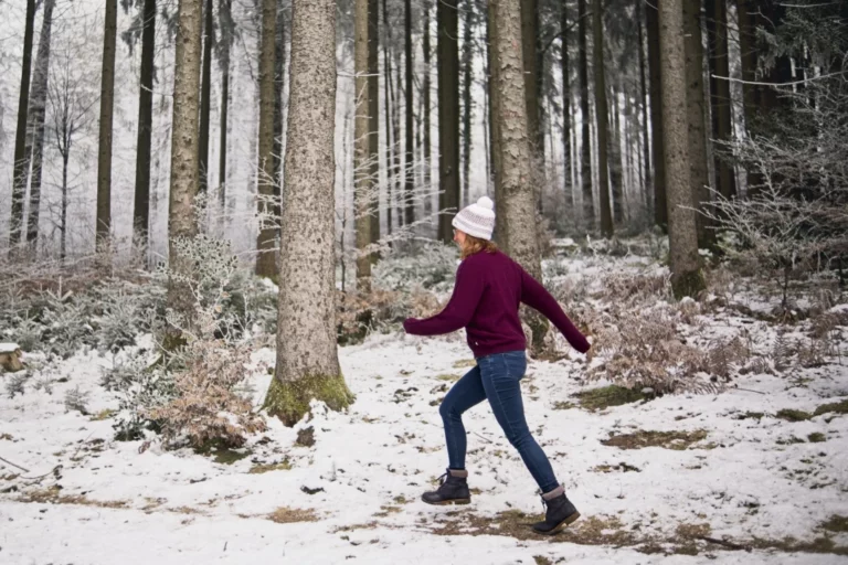 Eine Frau mit einer rosa Mütze und einem weinroten Pullover geht in einem verschneiten Wald spazieren. Sie trägt blaue Jeans und schwarze Stiefel. Die Bäume im Wald sind hoch und schlank, und der Boden ist mit Schnee bedeckt. Es gibt einige kleine Büsche am Boden, die auch mit Schnee bedeckt sind. Das Bild ist von der Seite aufgenommen und zeigt die Frau, wie sie durch den Wald geht.