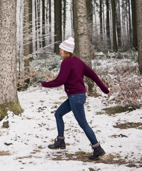 Eine Frau mit einer rosa Mütze und einem weinroten Pullover geht in einem verschneiten Wald spazieren. Sie trägt blaue Jeans und schwarze Stiefel. Die Bäume im Wald sind hoch und schlank, und der Boden ist mit Schnee bedeckt. Es gibt einige kleine Büsche am Boden, die auch mit Schnee bedeckt sind. Das Bild ist von der Seite aufgenommen und zeigt die Frau, wie sie durch den Wald geht.
