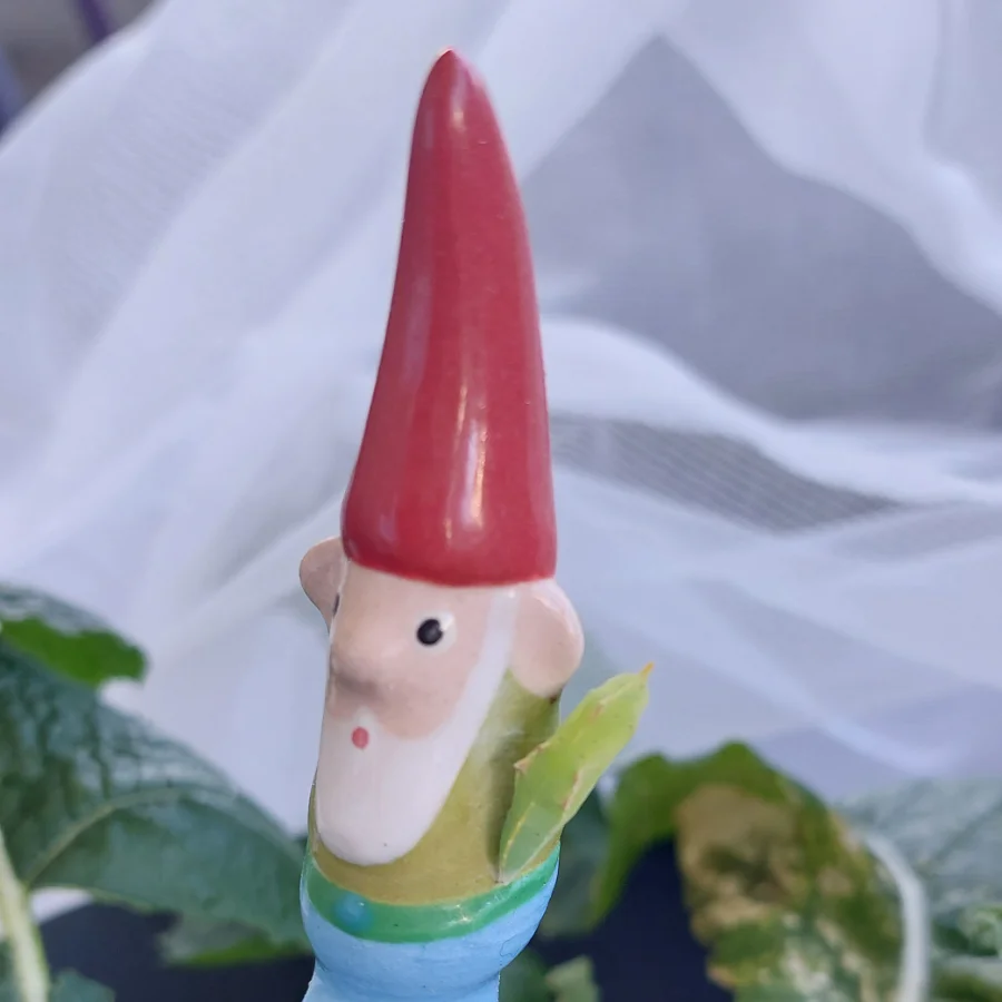 Detaillierte Nahaufnahme einer handbemalten Keramik-Gartenzwergfigur mit roter Mütze und weissem Bart, die ein kleines grünes Blattinsekt hält, umgeben von Pflanzen.