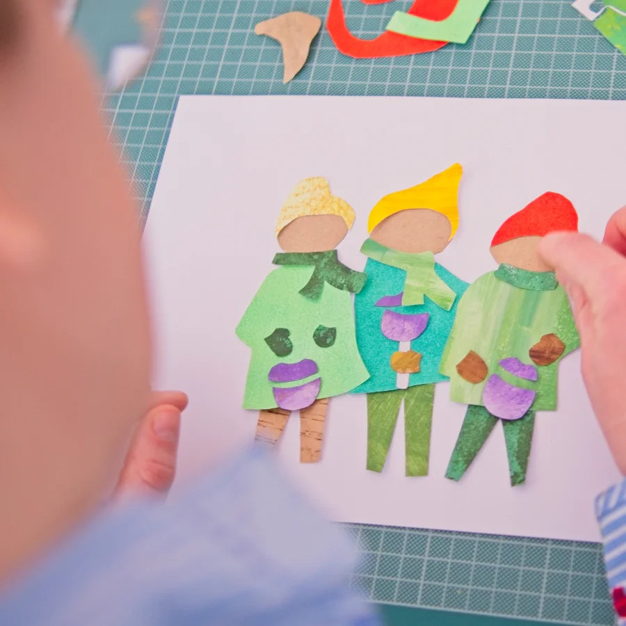 Person bastelt eine Collage aus buntem Papier mit drei kleinen Figuren auf einer grünen Schneidematte.
